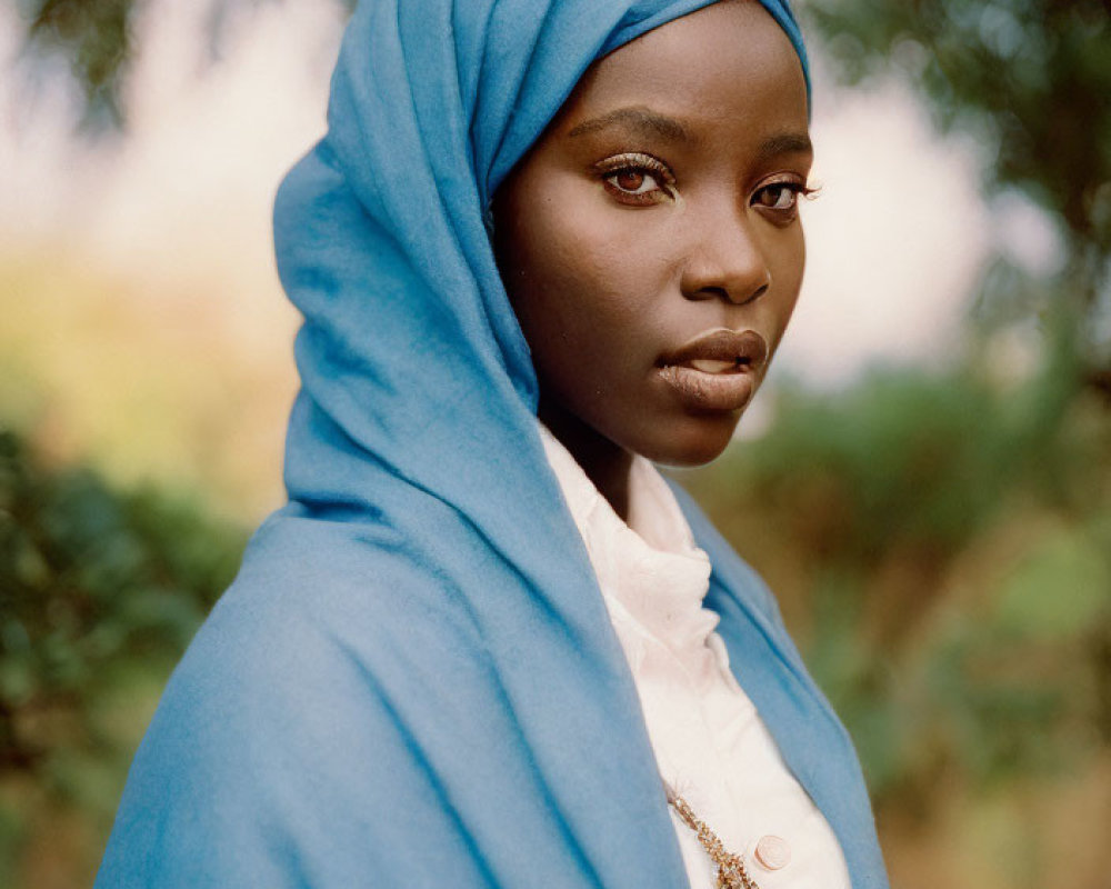 Dark-skinned woman in blue headscarf and white shirt with necklace against blurred nature backdrop