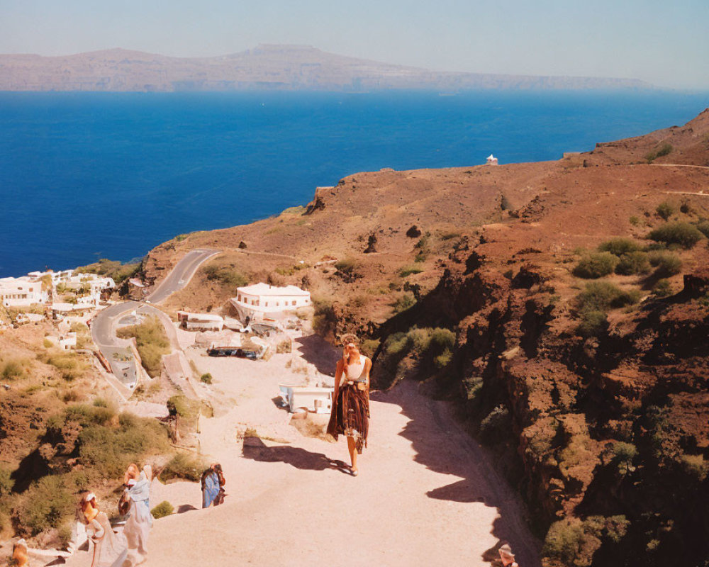 Person walking on dirt path with ocean view and white buildings in sunlight