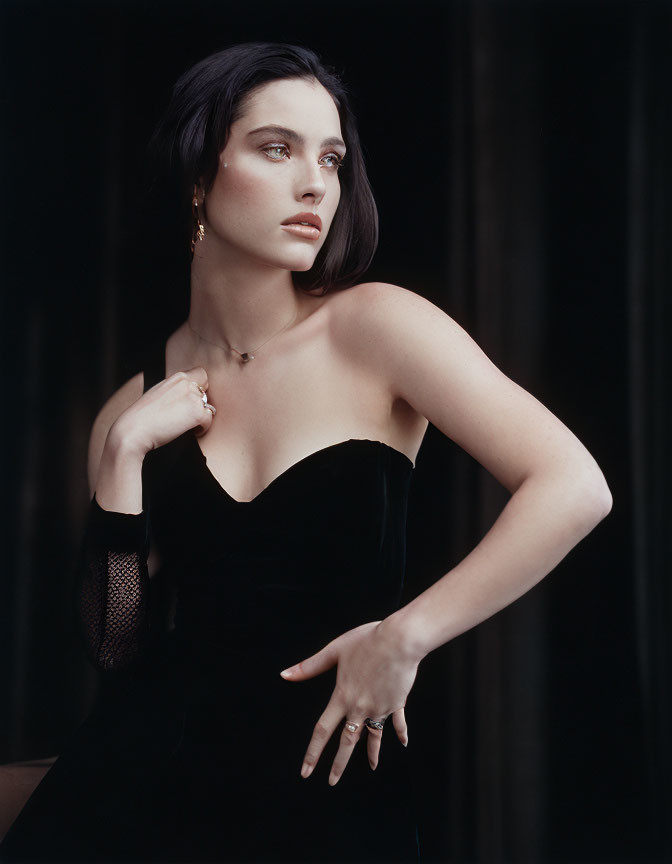 Dark-haired woman in black strapless dress and gloves poses against dark backdrop.