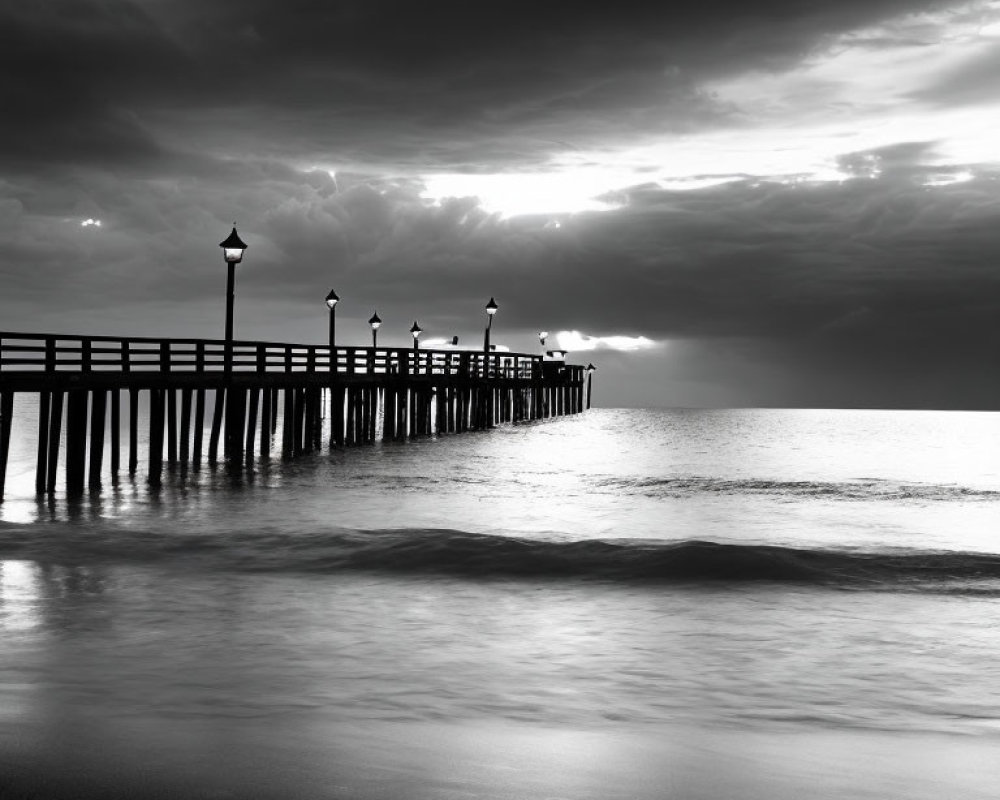 Monochrome image of calm sea with pier and lampposts under dramatic sky