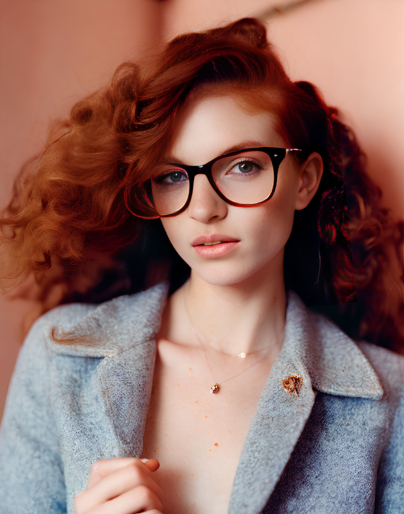 Wavy auburn hair woman in glasses with light blue coat on pink background