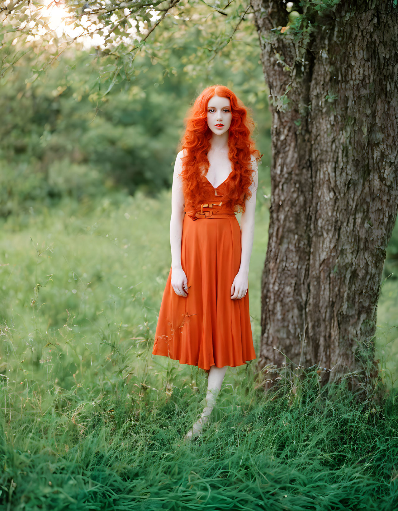 Red-haired person in orange dress near tree in lush green field with sunlight.