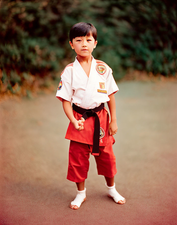 Child in karate uniform with black belt confidently standing on path with trees.