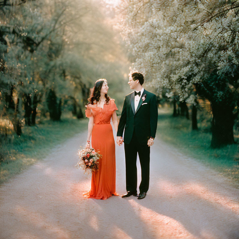 Formal couple holding hands on tree-lined path with woman in orange dress and bouquet in serene setting