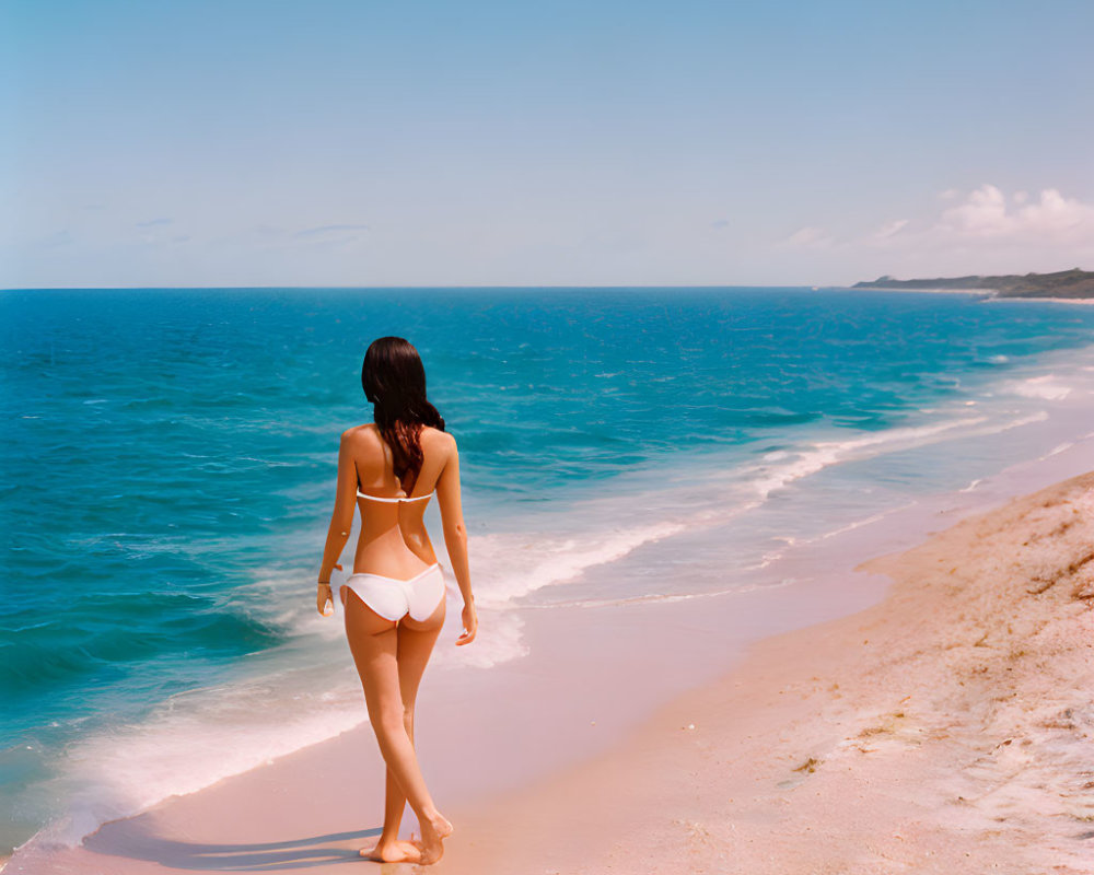 Person in white bikini on sandy beach gazing at calm blue ocean