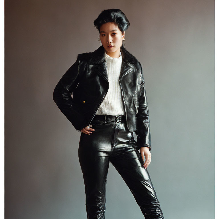 Fashionable person in black leather jacket and pants with white sweater posing confidently.