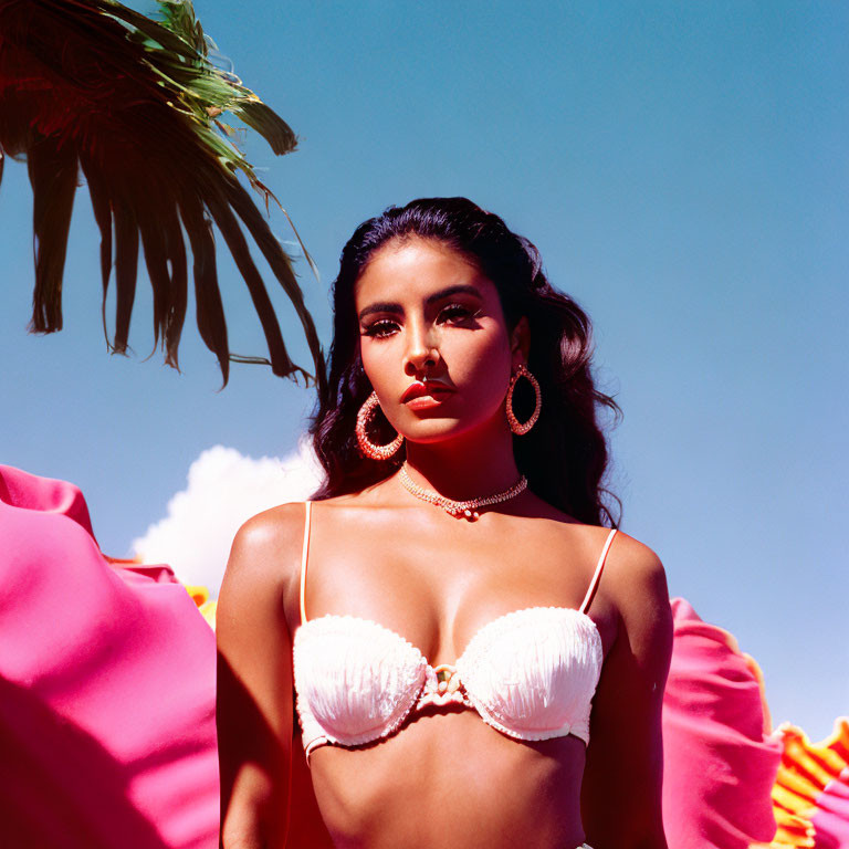 Woman in white bikini top and gold jewelry under blue sky with palm leaf and pink fabric.