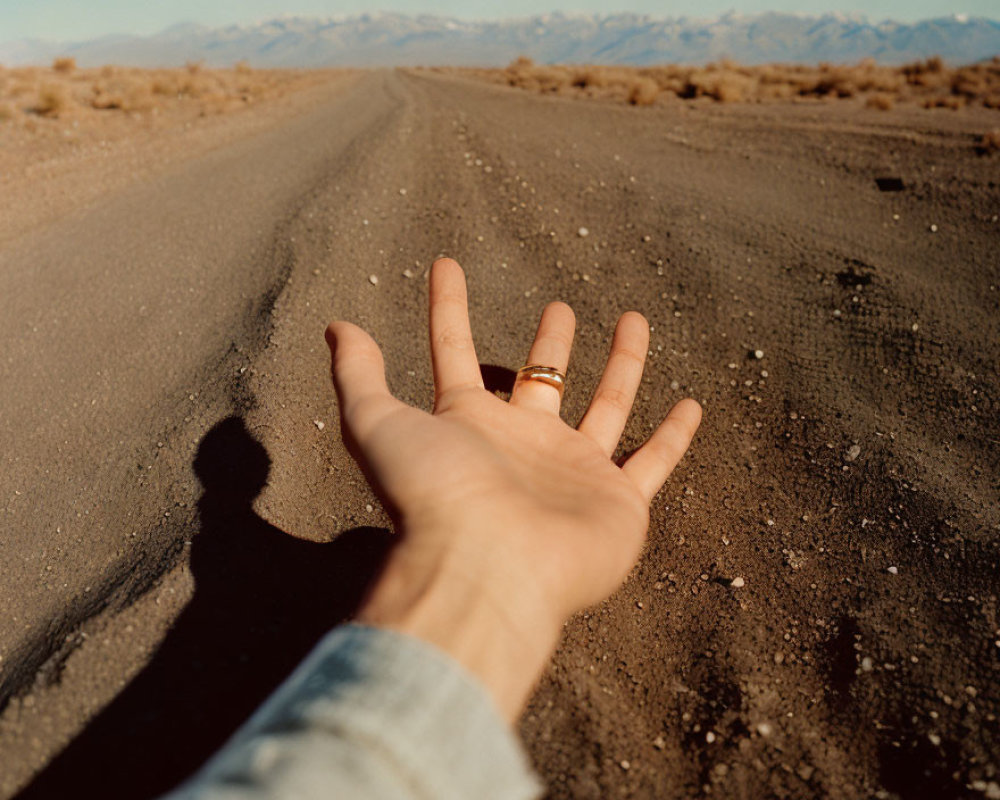 Hand with ring on finger on dirt road with shadow and mountains.