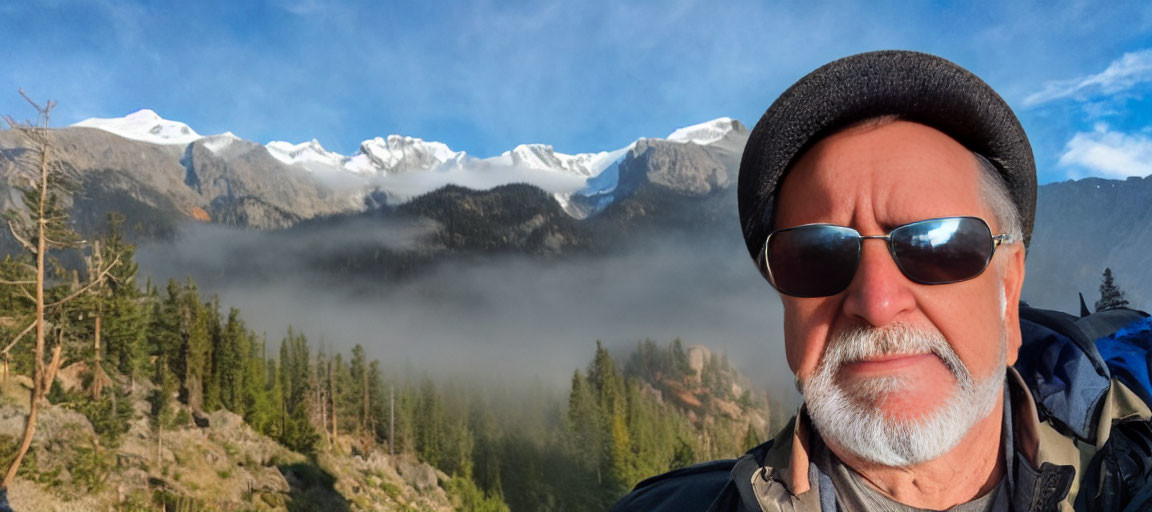 Man in Sunglasses and Hat Posing in Front of Misty Snow-Capped Mountains