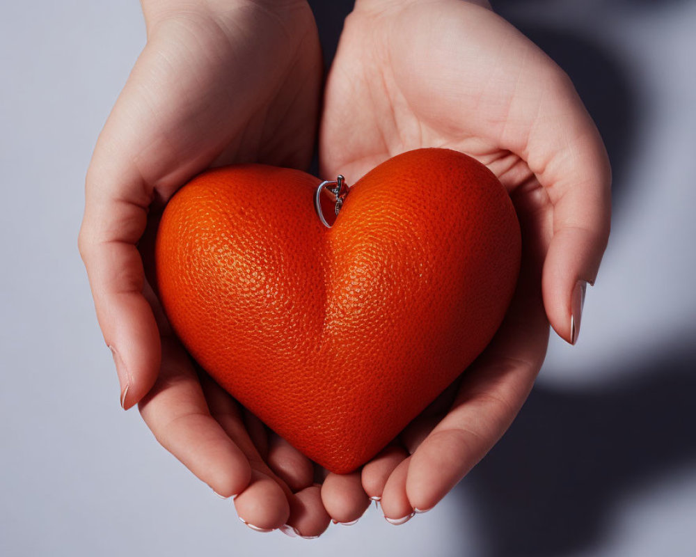 Hands holding heart-shaped orange object with loop on pale background
