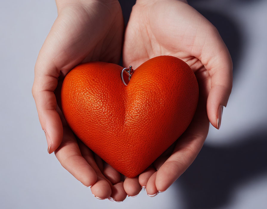 Hands holding heart-shaped orange object with loop on pale background