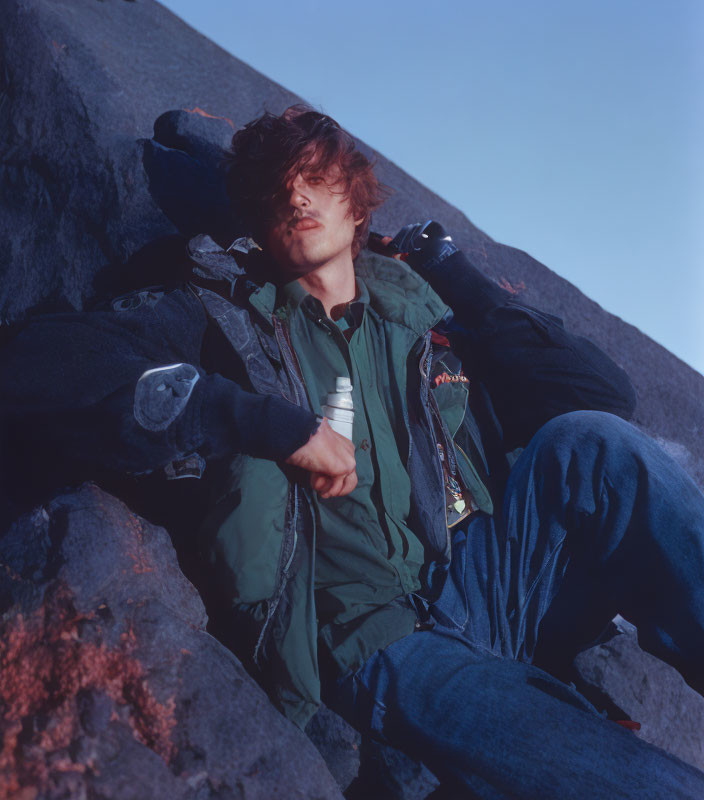Messy-haired person in green jacket on rocky terrain with water bottle