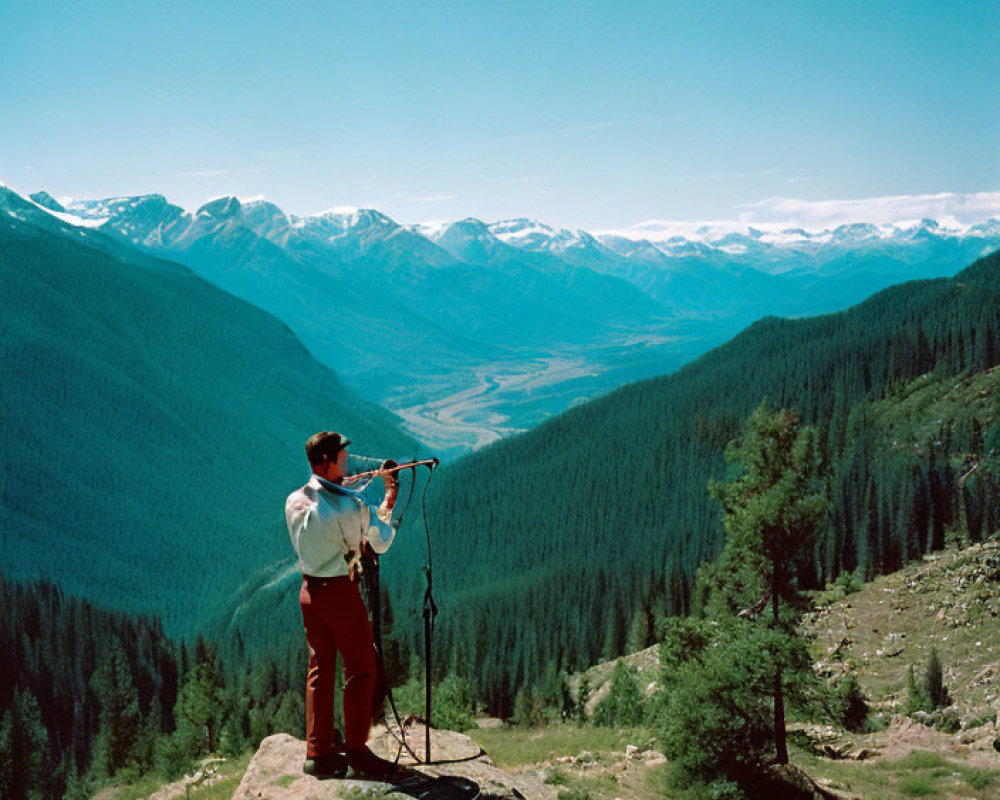 Person with binoculars on forested mountain trail overlooking snow-capped peaks