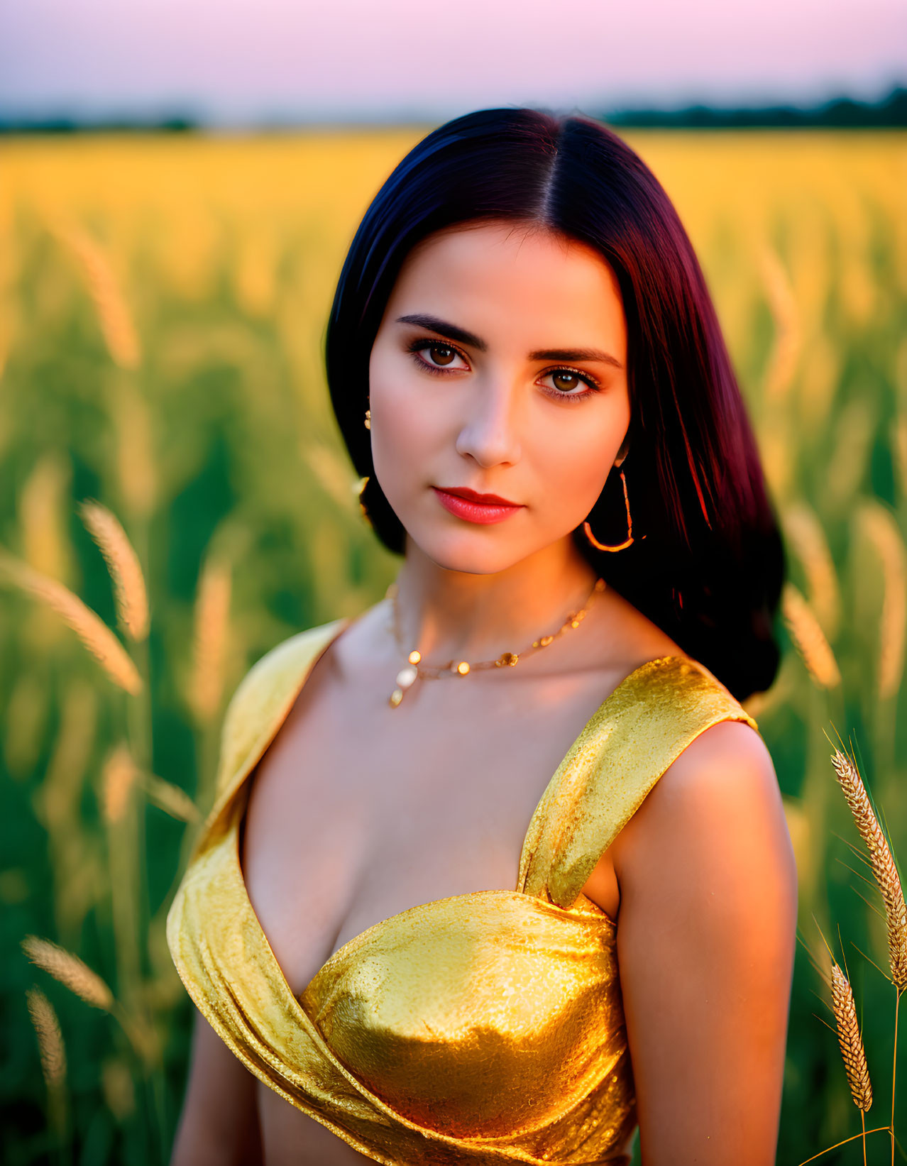 Dark-haired woman in golden top posing in sunset-lit field.