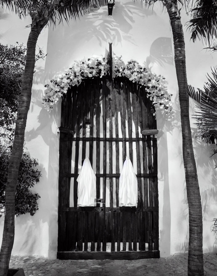 Monochrome image: Ornate wooden gate with flowers, palm trees, and white cloths