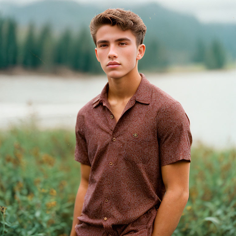 Young man in brown shirt posing by lake and mountains.
