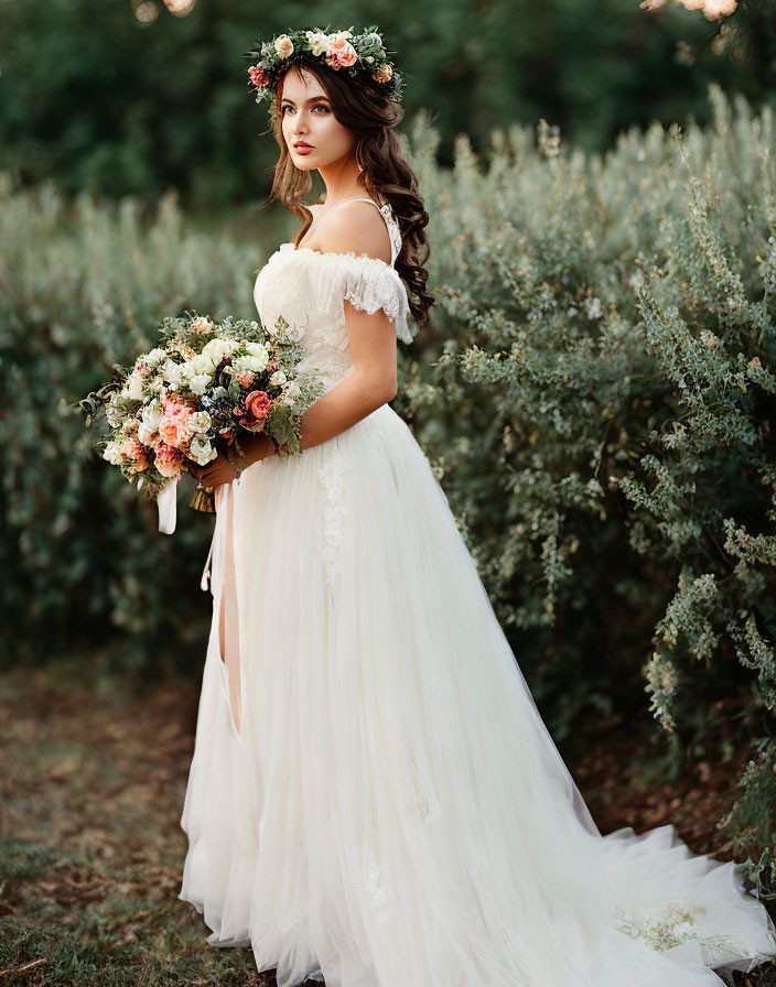 Bride in white off-shoulder gown with floral crown holding bouquet in greenery