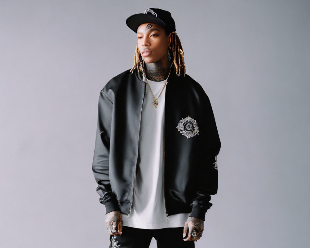 Fashionable person in black bomber jacket, white tee, cap, and jewelry on grey background