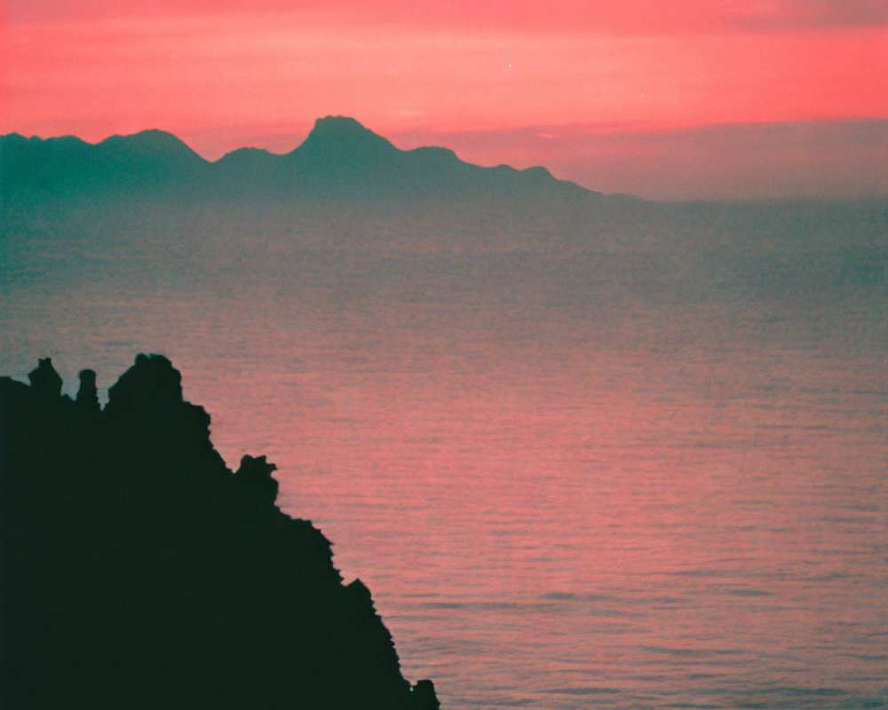 Colorful pink and red sunset over calm sea with silhouetted figures on cliff and distant mountains