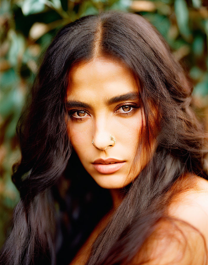 Woman with long dark hair and nose ring in intense gaze against green foliage.