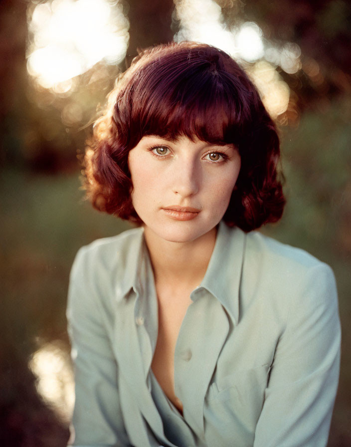 Woman with short curly brown hair in light blue shirt gazes at camera in serene setting