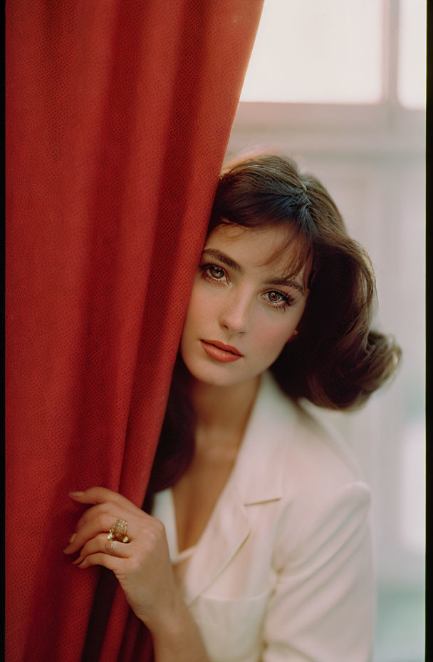 Woman with dark hair and white blouse peeking from red curtain
