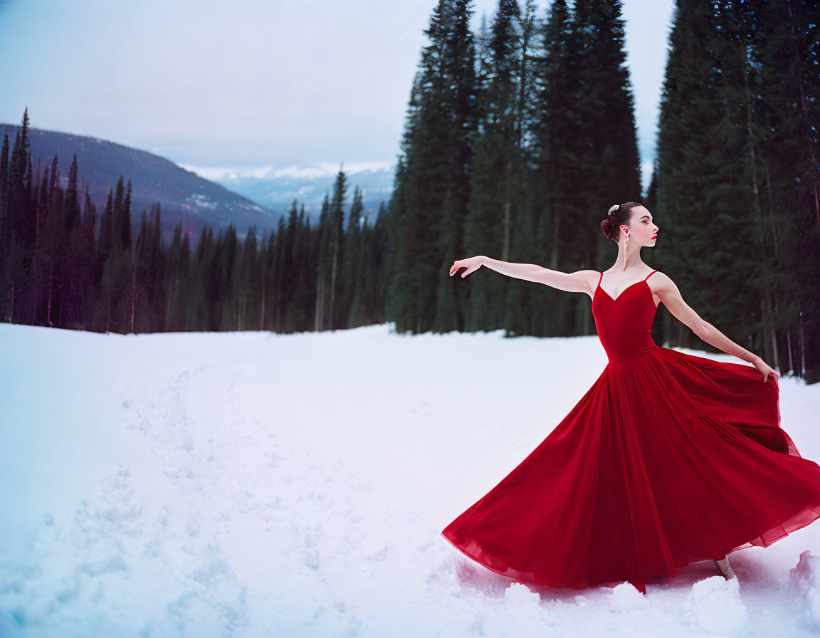 Woman in red dress posing in snowy forest landscape with pine trees and mountain.