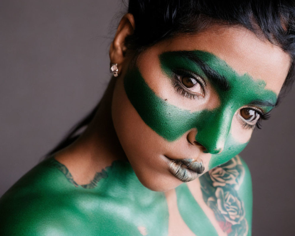 Person with green paint on face and body staring at camera with shoulder tattoo.