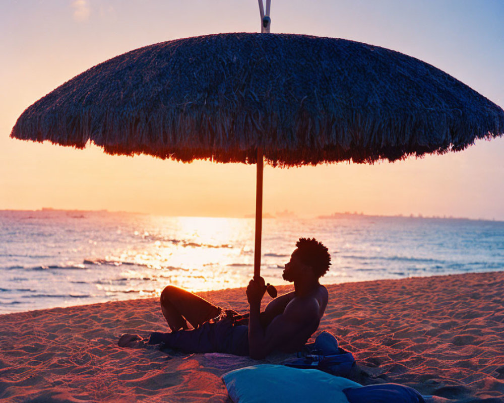 Person relaxing under thatched beach umbrella at sunset with ocean view