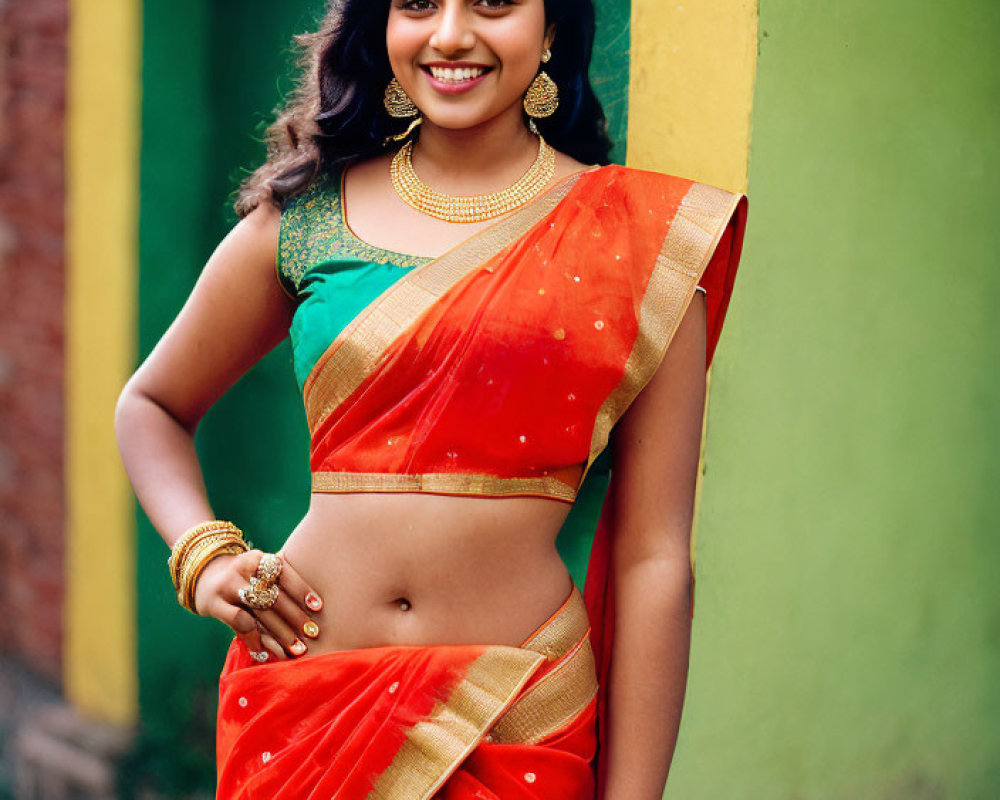 Smiling woman in red saree with gold jewelry against yellow and green wall