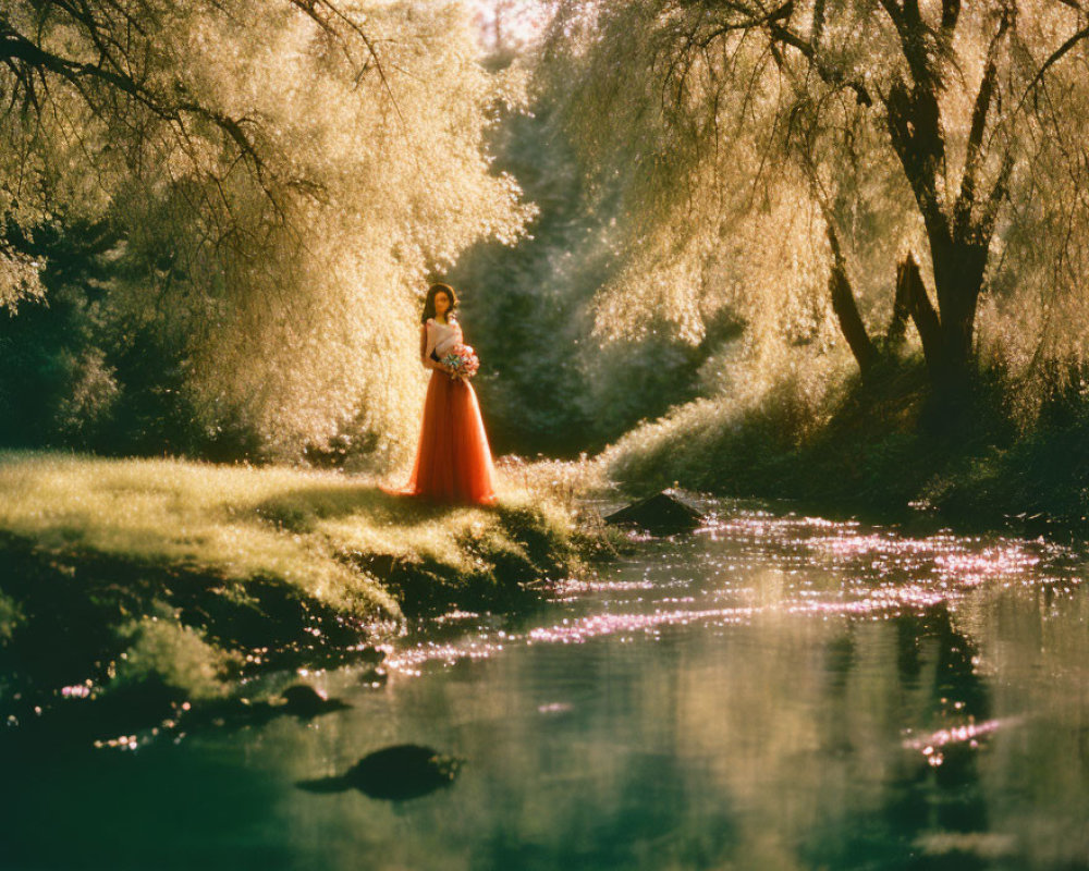 Woman in flowing orange gown by tranquil stream under sunlit willow tree
