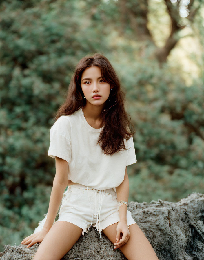 Serene young woman sitting on rock in casual attire surrounded by greenery