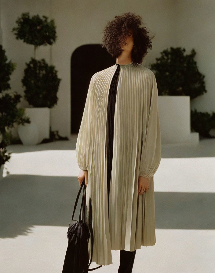 Person in cream pleated cape holding black bag in sunlit courtyard
