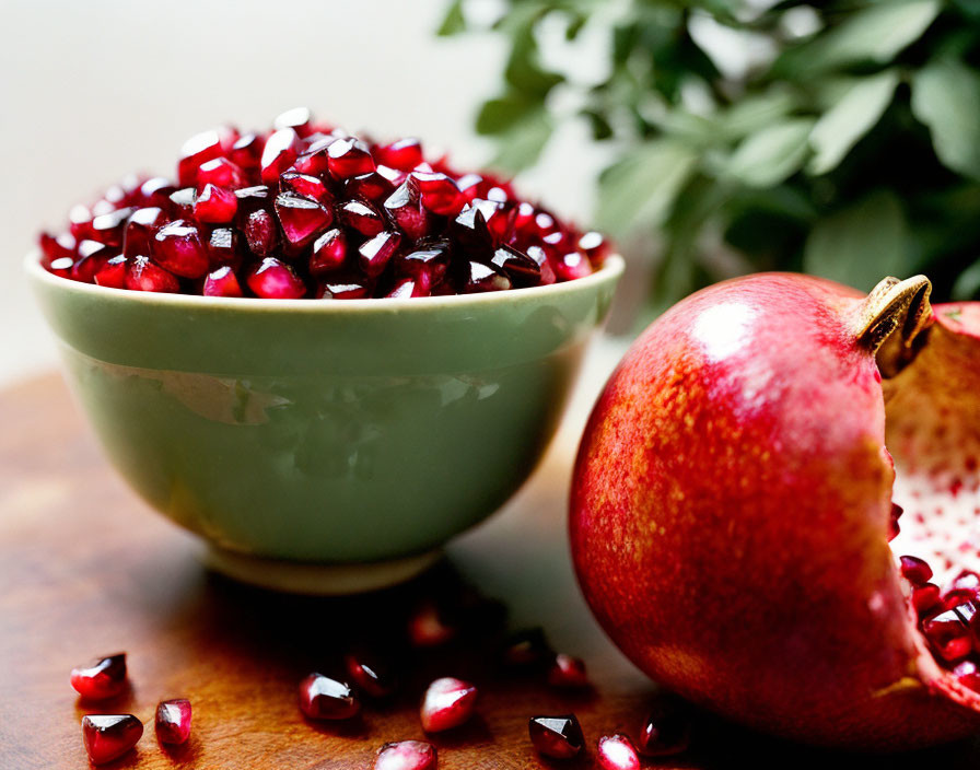 Green bowl with pomegranate seeds, whole and opened fruit on wooden surface
