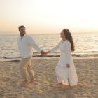 Couple holding hands on beach at sunset in white attire.