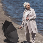 Elderly woman with white hair holding black umbrella by lake in trench coat