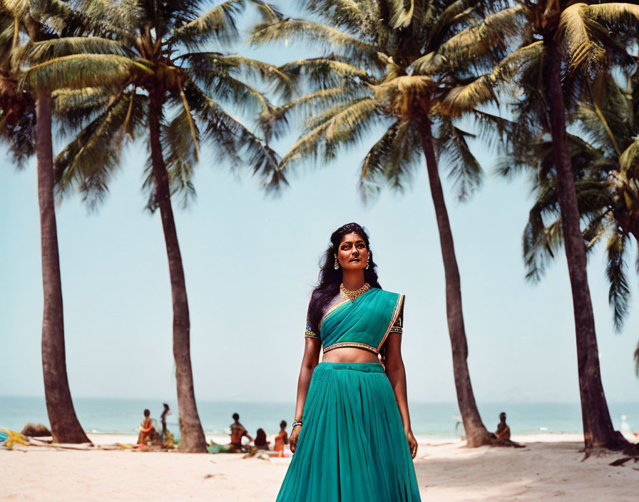 Woman in teal saree on sunny beach with palm trees and people.