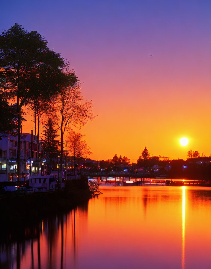 Vibrant sunset reflected in calm river with silhouetted trees and distant city lights