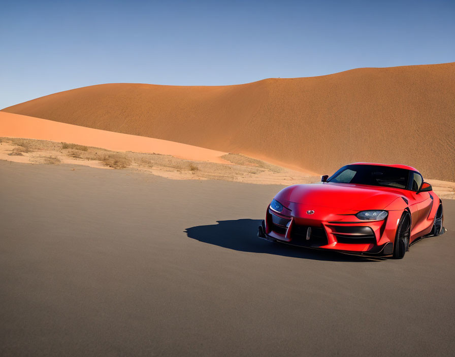 Red sports car parked in vast desert landscape with smooth sand dunes under clear blue sky