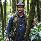 Bearded man in hat, vest, green shirt, with rifle in lush forest