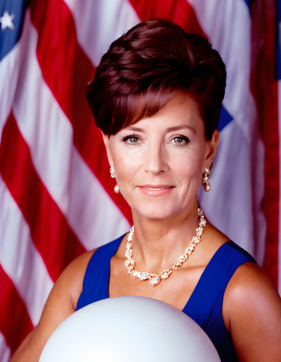 Smiling woman with short brown hair in blue dress and pearl necklace in front of American flag