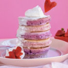 Colorful stack of pancakes with pink and blue icing, cream swirl, and strawberries on pink background