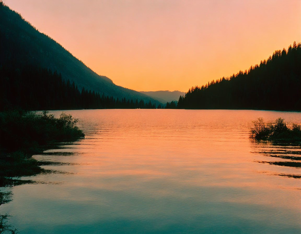 Tranquil sunset reflected in serene lake with forested hills.