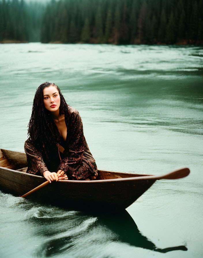 Woman in patterned outfit in wooden canoe on serene river with lush green trees and moody sky.