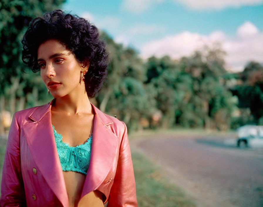 Curly-haired woman in pink jacket by vintage car and trees.