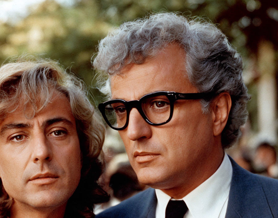 Two mature men with blond and gray hair, one wearing black glasses, looking serious.