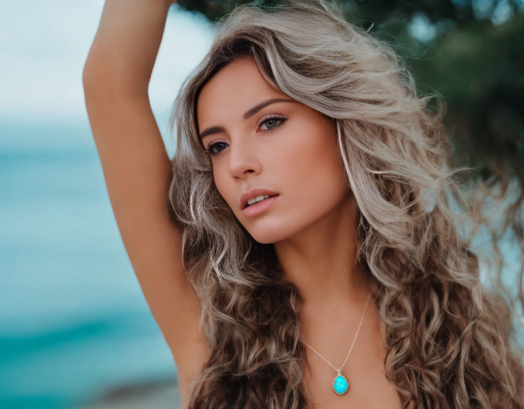 Wavy-haired woman in turquoise necklace gazes at seaside.