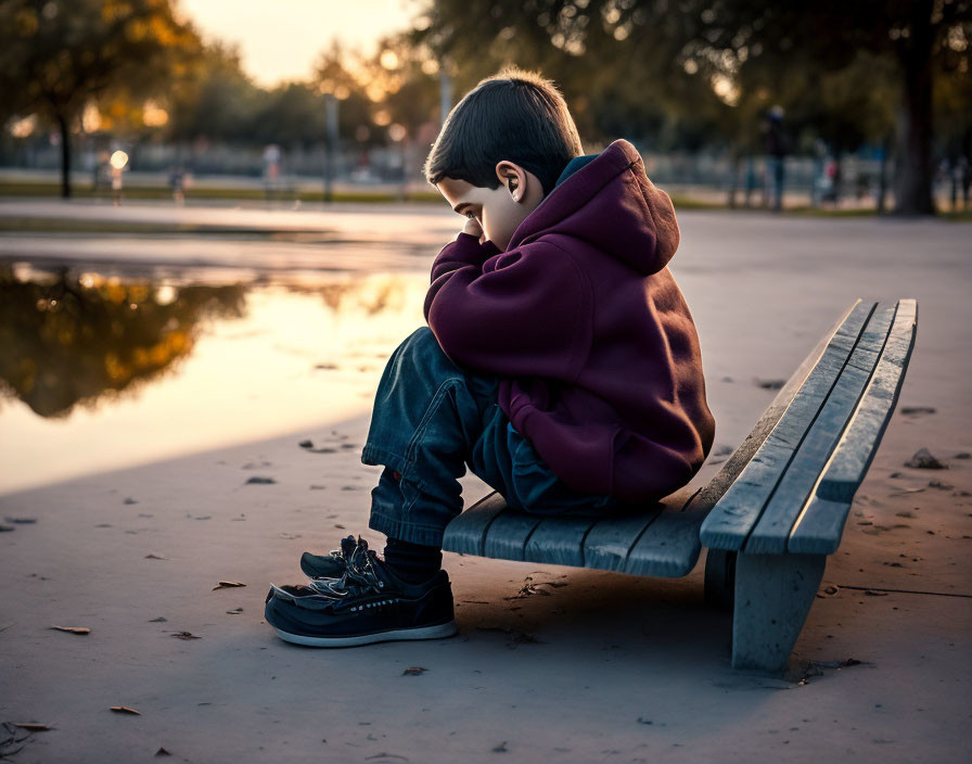 Young person sitting on park bench near water body at sunset