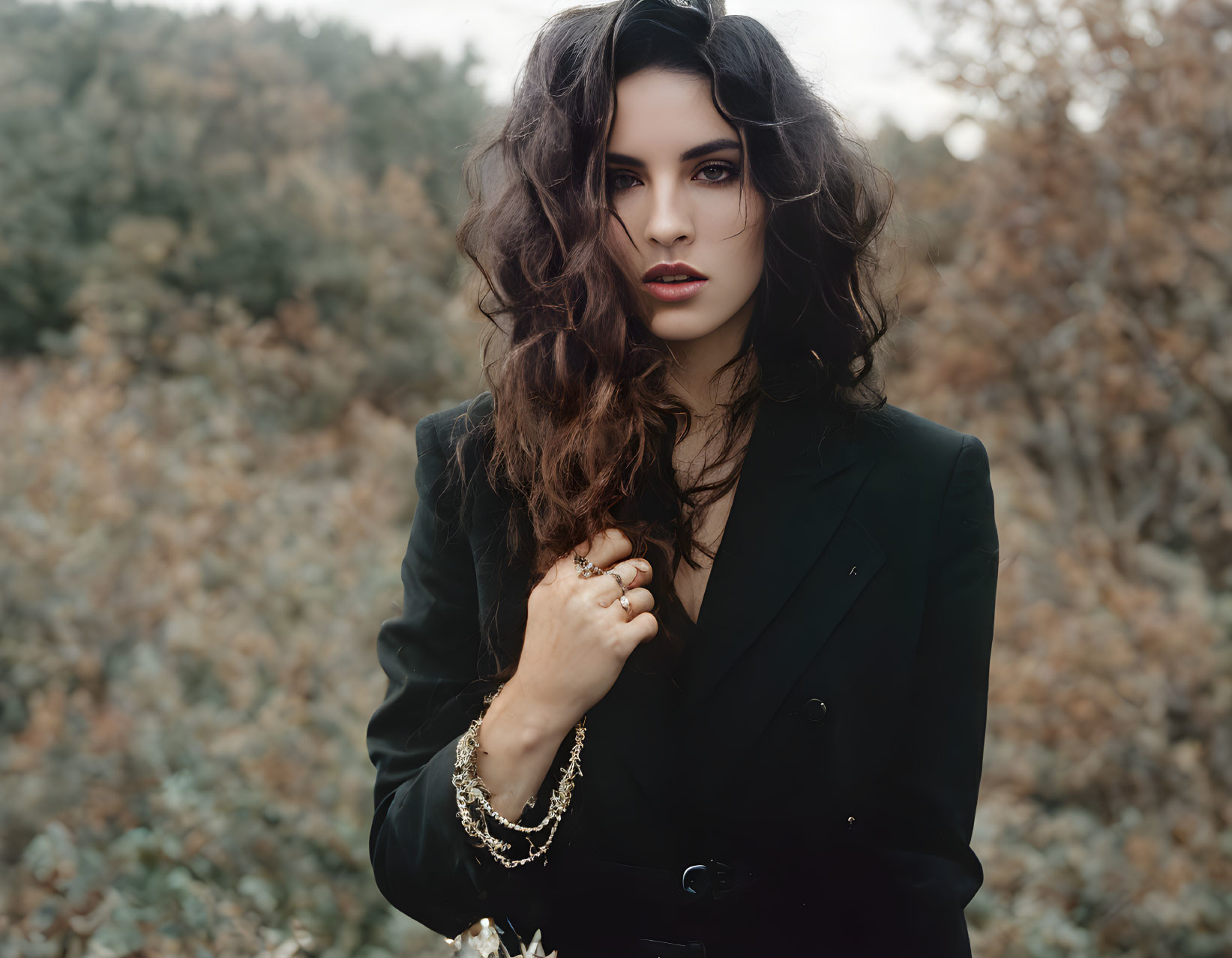 Curly-haired woman in black blazer with bold makeup against natural backdrop