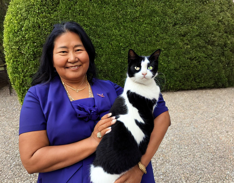 Smiling woman in blue dress with black and white cat by green hedge